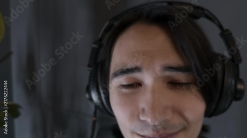 Intense young Asian male podcast host finishing emotional true crime audio storytelling session taking off black headphones near condenser microphone in dark modern digital media studio tracking shot