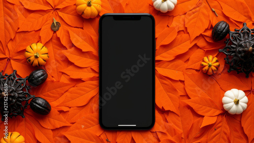 Black smartphone centered on a vivid orange Halloween flat lay with autumn leaves and festive decorations providing a bold seasonal mobile mockup for promotions apps and campaigns