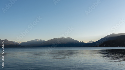 Фотография Calm lake water stretches toward distant mountains under a clear blue sky with soft lighting, reflecting the peaceful morning landscape