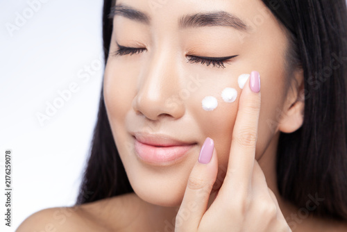 Beauty portrait of calm woman with closed eyes touching cream on cheek with her finger while enjoying skincare routine isolated over gray background