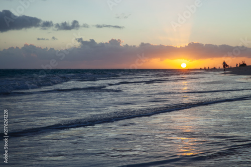 Golden sunset glows on the horizon as gentle waves wash a reflective shoreline. Silhouetted figures stroll the sandy beach, creating a peaceful, romantic and contemplative coastal scene at dusk