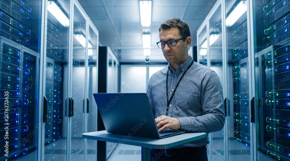 Fototapeta premium Man engineer working with laptop in data center server room. IT specialist checking network equipment in modern datacenter. Cloud computing, cybersecurity and digital technology concept