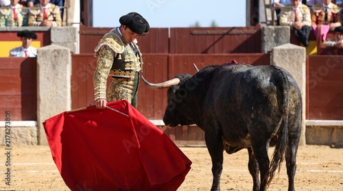 Matador in traditional gold embroidered costume holding red cape facing black bull in bullfighting arena. Spanish bullfight corrida spectacle with