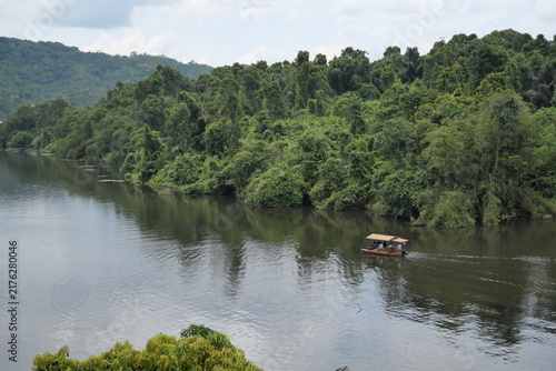 A tranquil tropical lake surrounded by dense lush rainforest and green hills, with a small traditional boat cruising across calm water.