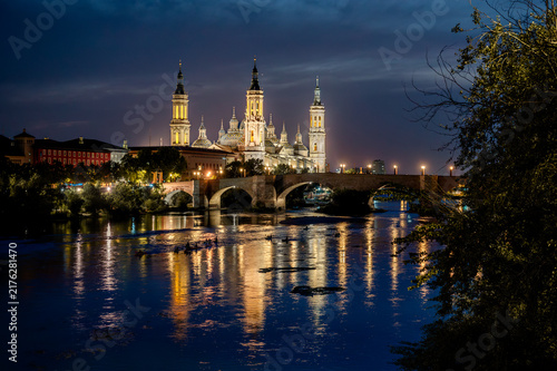Obraz na plátne The illuminated Basilica del Pilar reflects in the calm Ebro river beside the Puente de Piedra stone bridge at blue hour in Zaragoza Spain an iconic cityscape of the Aragon landmark at night