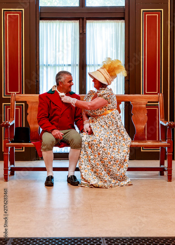 Obraz na plátně A man and a woman dressed in 19th-century historical attire sit on a wooden bench, where the woman affectionately adjusts the man's cravat during a historical reenactment or themed event