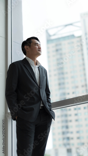 Fotografie Confident Asian businessman in suit standing near glass window looking out at cityscape