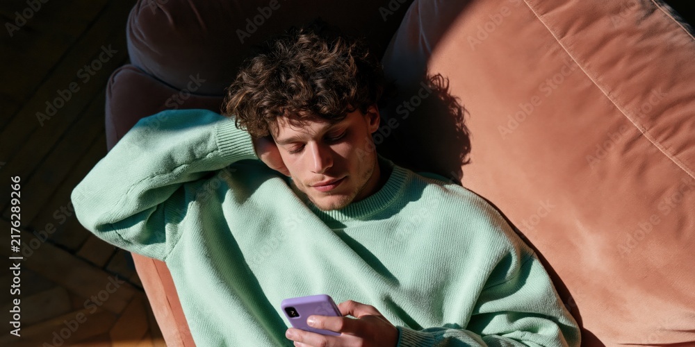 Obraz premium Young man lying on terracotta couch checking smartphone with arm behind head in mint sweater warm window light for social media lifestyle visual