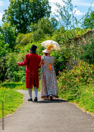 Photography A couple in historical Regency-era costumes, featuring a red frock coat, top hat, and floral gown, walk arm-in-arm along a garden path while participating in a period reenactment event on a sunny day