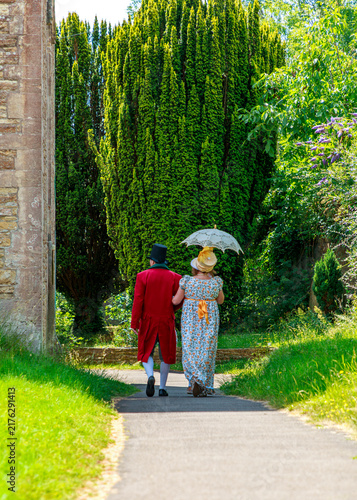 Fotografie A couple dressed in Regency-era historical attire walk arm-in-arm along a garden path, likely participating in a historical reenactment event