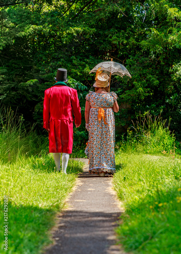 Canvas Print A man in a historical red tailcoat and a woman in a period floral dress stroll along a garden path, likely participating in a Regency or Victorian-era costume reenactment event in nature