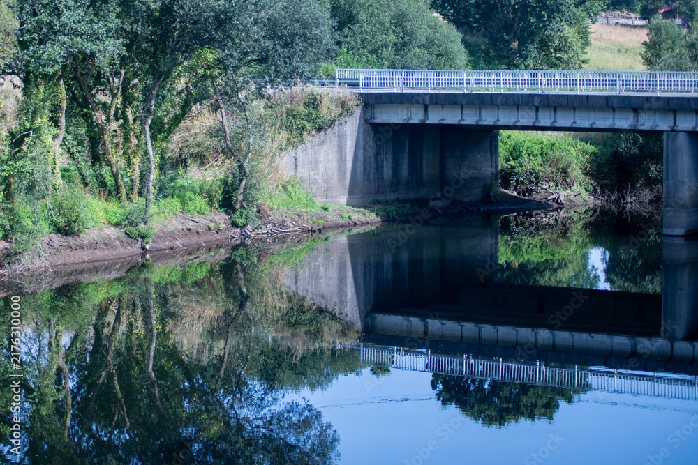 Fototapeta premium A concrete road bridge over a calm river reflecting symmetrically in the still water surrounded by green trees.