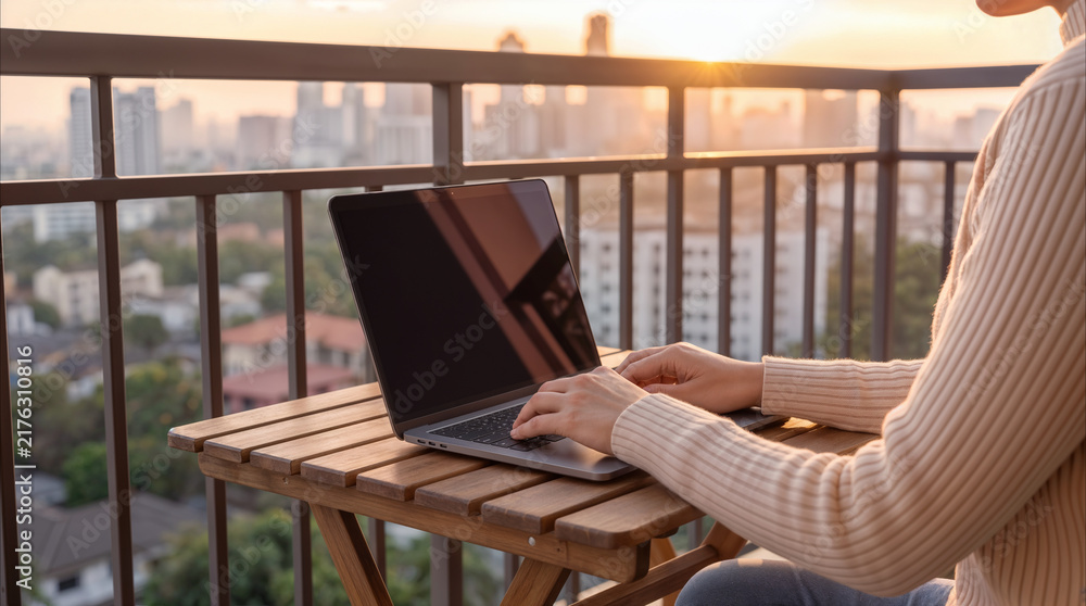 Obraz premium Freelance professional typing on laptop at sunset on apartment balcony. Remote work from home office with city skyline view.