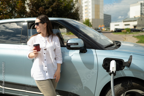 Beautiful woman with coffee leaning on electric car during charging