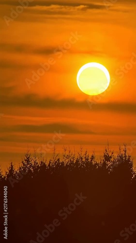 Partial solar eclipse over a treeline silhouette at golden sunset. sun briefly occluded then reemerges through hazy orange clouds.