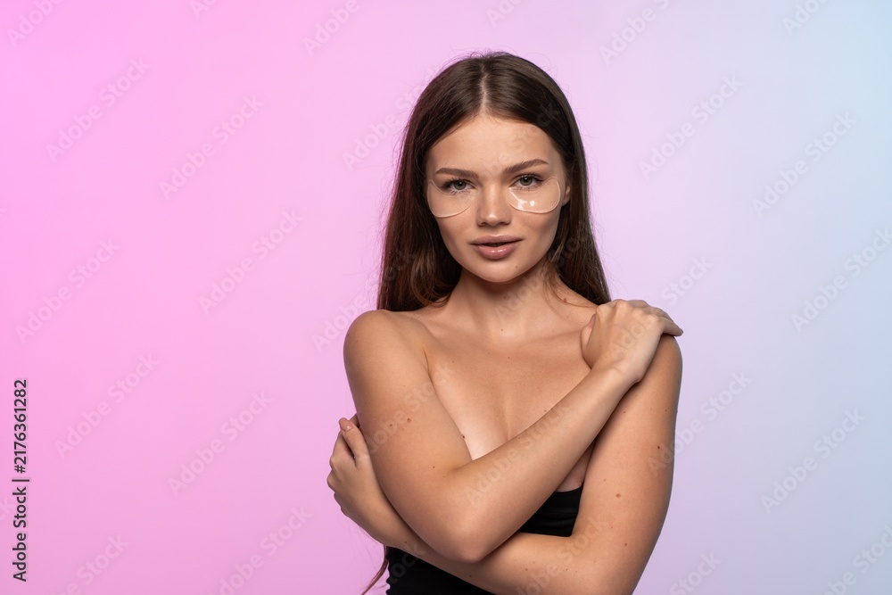 Fototapeta premium Young woman applying under eye patches for skincare routine, embracing beauty and self-care on gradient background