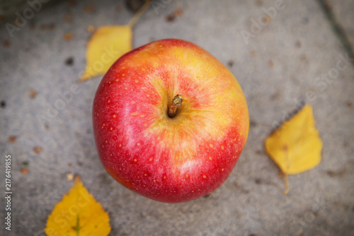 A red apple lies on the asphalt next to some yellow leaves.