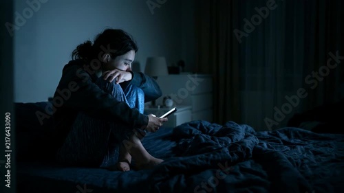 Sad woman sitting on bed at night looking at phone. Heartbroken girl feeling lonely and depressed due to betrayal. Conflict in marriage and relationship crisis.