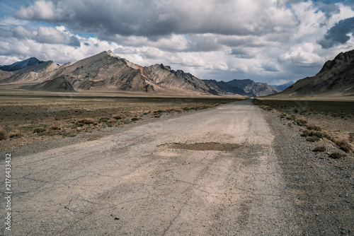 View of the Pamir Mountains with a rugged dirt road stretching through the arid landscape under a dramatic cloudy sky in Jirgatol, Gorno-Badakhshan Auto, Tajikistan.