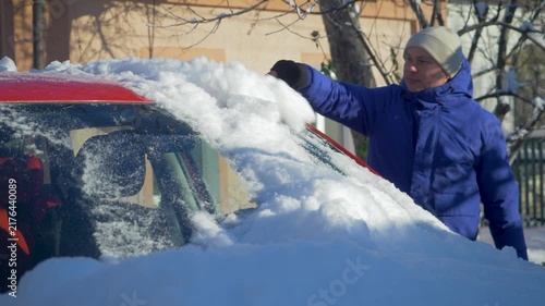Man uses snow brush to clear front glass of red car covered by heavy winter precipitation. Frost and deep snow limit visibility. Severe cold creates demanding conditions for morning travel