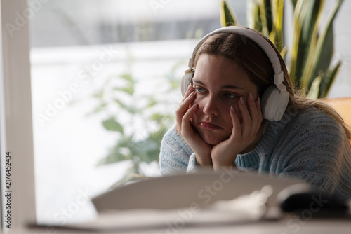 Tired young Caucasian woman wearing headphones resting her face in her hands while looking at a laptop screen, feeling bored, exhausted, or frustrated while working or studying from home