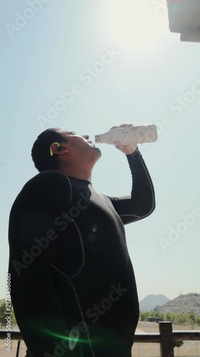 Thirsty male athlete in a wetsuit drinking water from a reusable bottle on a hot sunny day to stay hydrated during his workout and rehydrate his body