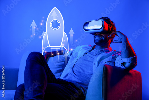 Fotografie A dynamic scene capturing a joyful entrepreneur using virtual reality, envisioning a future business success amid vibrant blue lighting with imaginative rocket graphics