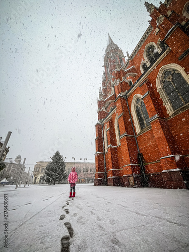 Fotografía Snow falls on a child standing near a tall red brick building in a winter scene