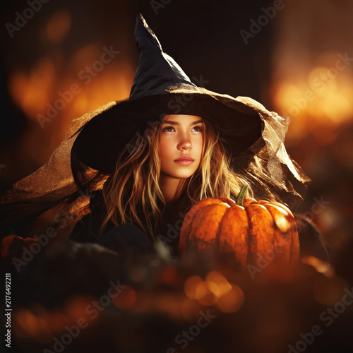 Young girl wearing witch hat holding pumpkin in warm autumn glowing light with magical atmosphere