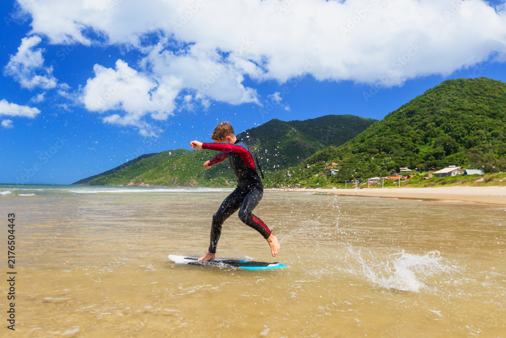 Fototapeta premium Young boy skimboarding in shallow water on a sunny beach day. Wearing wetsuit, balancing on board with determination and focus. Perfect image for themes of childhood adventure, active lifestyle