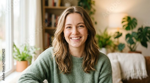 Smiling young woman with wavy brown hair wearing sage green sweater, cheerful head-and-shoulders portrait in cozy living room with houseplants and bookshelf background