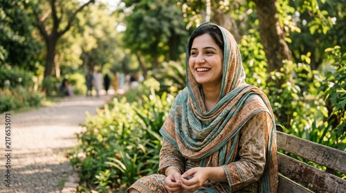 Smiling Pakistani woman in traditional patterned scarf sitting on park bench surrounded by lush greenery on sunny day candid natural portrait