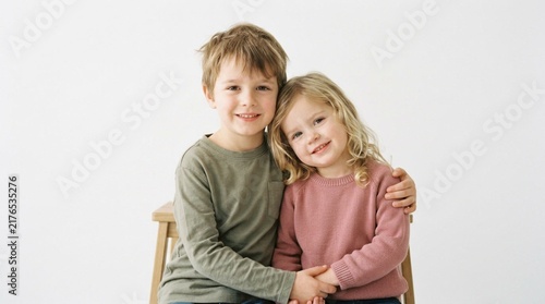 Smiling young brother and sister embracing in cozy sweaters against clean white studio backdrop with soft diffused lighting, happy childhood portrait