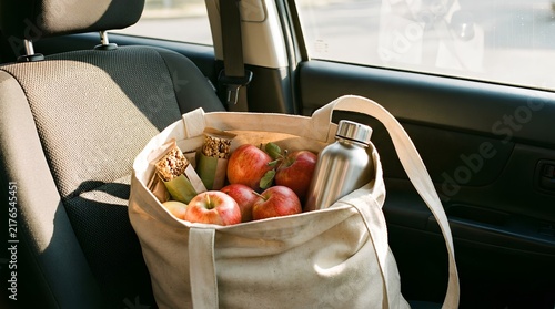 Canvas tote bag filled with fresh red apples, granola snack bars, and a stainless steel reusable water bottle resting on a gray fabric car passenger seat during a sunny road trip