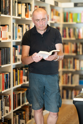 фотографія Focused middle-aged guy browsing through literature volumes on shelves in cozy b