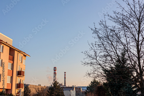 Quadro su tela Apartment Building and Industrial Smokestacks at Sunset With Bare Trees in Urban