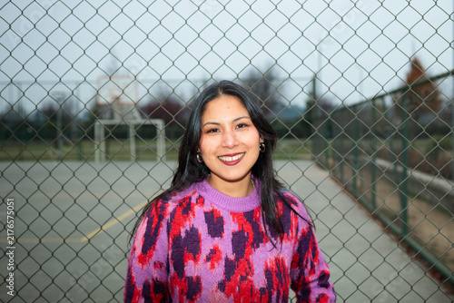 Cheerful young latina woman smiling in front of a wire fence at an outdoor sports court