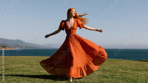 Woman in Long Orange Dress Dancing on Grass by the Sea