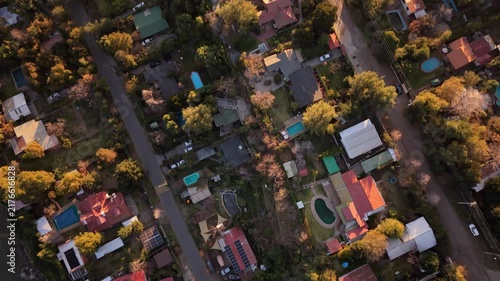 Top down nadir shot of a suburban residential neighborhood with colorful rooftops, backyard swimming pools, and golden autumn trees lining quiet streets at sunset