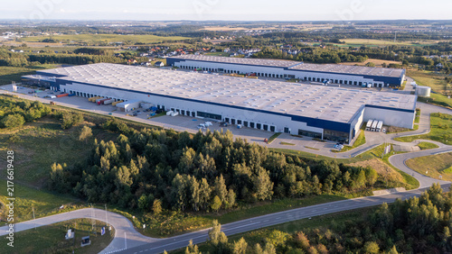Aerial view of a large logistics warehouse and distribution center with multiple loading docks and parked trucks surrounded by green fields and trees in Gdansk, Poland.