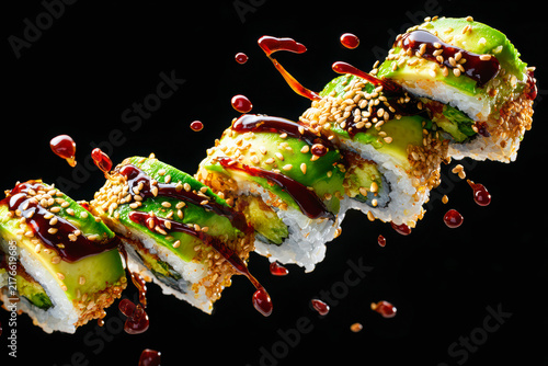Avocado topped uramaki sushi rolls tumble diagonally through the air against a black background as dark sauce drips and sesame seeds scatter around them.