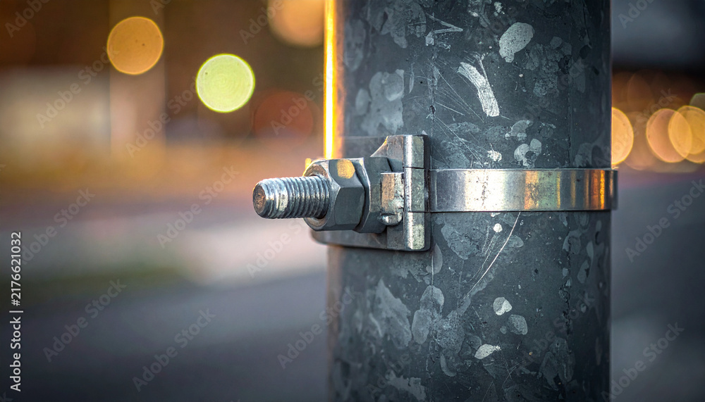 Fototapeta premium Streetlight Clamp Detail: A close-up shot reveals the sturdy metal clamp securing a bolt onto a weathered streetlight pole, with blurred city lights in the background creating a bokeh effect.