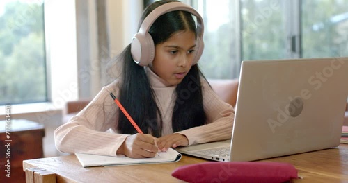 Indian schoolgirl with headset at home writing in notebook, glancing at silver laptop, raising hand