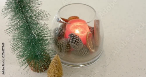 When breeze sweeps speckled countertop, red candle flame is flickering, casting glow on pine cones