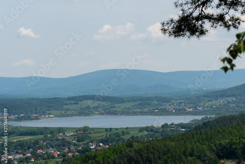 Wide panoramic view of a lake surrounded by villages, green fields and forested mountain slopes in Poland. Peaceful summer countryside landscape. Concept of eco tourism and scenic travel.
