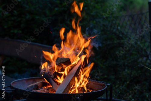 Bright orange flames burning stacked firewood in a metal fire pit against a dark evening background. Concept of outdoor cooking, summer barbecue, warmth, energy and campfire safety.