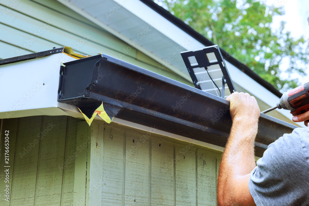 Obraz premium Contractor attaches gutter to edge of building while using drill with house with green siding.