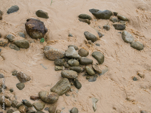 Fotografie Natural hazards during the rainy season river surges and mudslides rapidly flowing muddy brown water rushes past dark scattered rocks weeds and trees within the stream or river