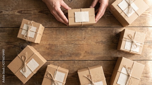 Hands arranging small brown gift boxes tied with twine and blank labels on a rustic wooden table