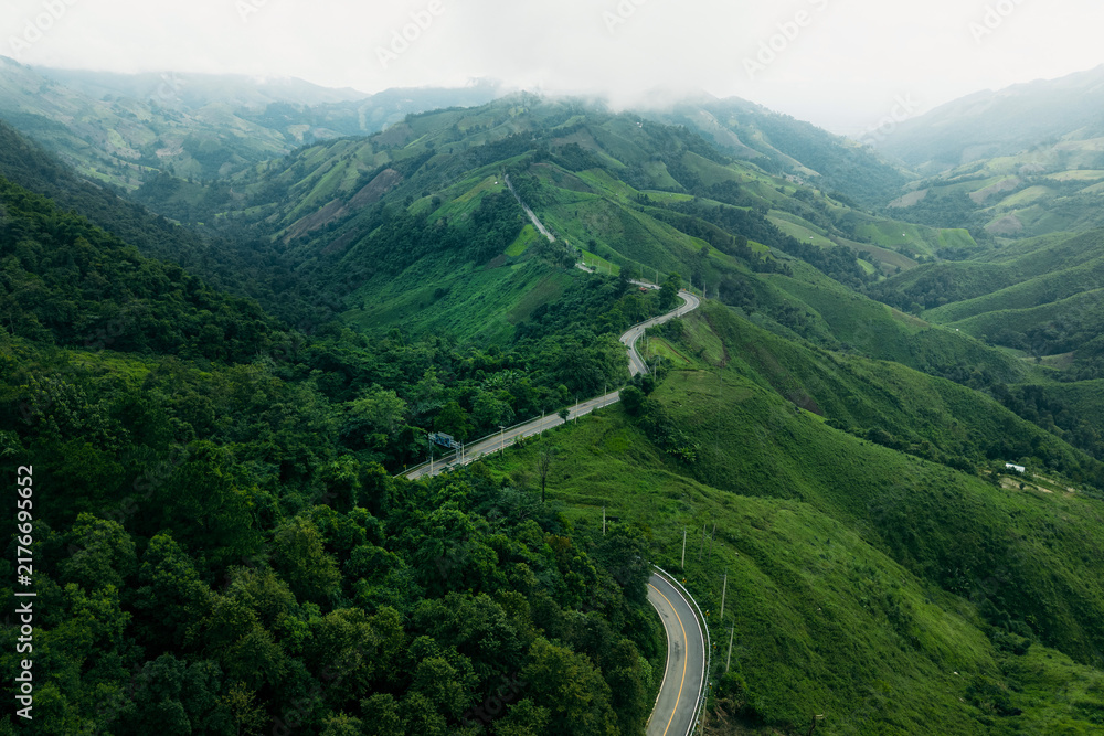 Obraz premium Aerial view of a winding asphalt road cutting through lush green mountains and tropical rainforest.
